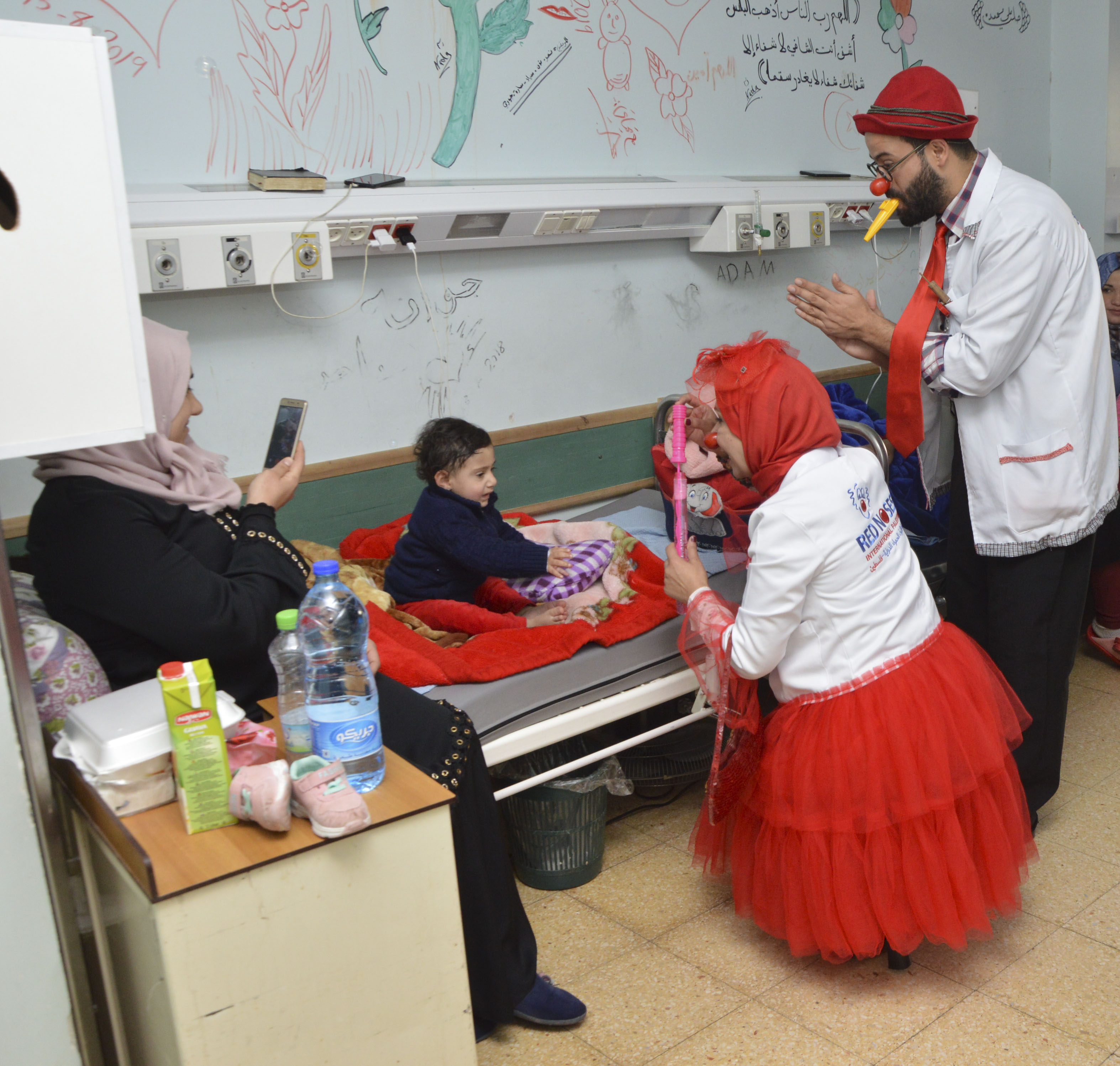 Two RED NOSES clowns, male and female, play with baloons in their hand with a small child on his hospital bed as his mother is taking a picture with her phone. a